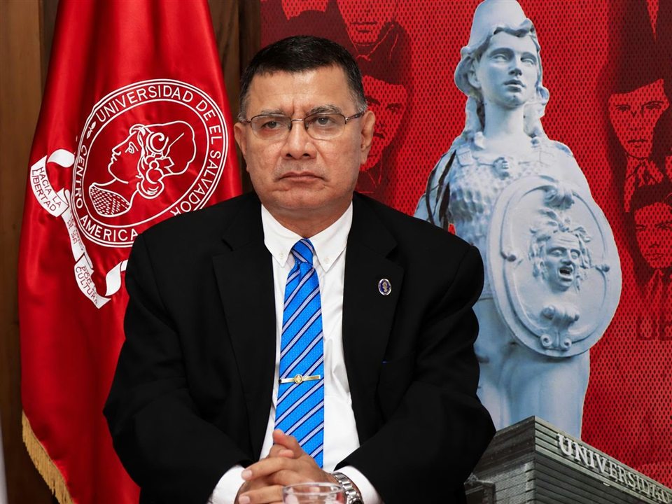 Retrato institucional de un hombre de traje junto a la bandera de la Universidad de El Salvador